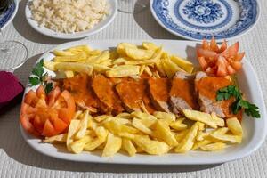 A plate of veal stew in a rich tomato sauce, served with crispy fries, is presented on a table at a traditional Alentejo restaurant. The rustic setting and hearty dish create a inviting atmosphere. photo