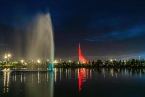 UZBEKISTAN, TASHKENT - SEPTEMBER 15, 2023 Illuminated monument of independence in the form of a stele with a Humo bird and fountain in the New Uzbekistan park at nighttime in autumn. photo