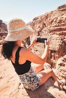 maravilloso caucásico mujer turista sentar en punto de vista en petra antiguo ciudad terminado tesorería o al-khazneh tomar teléfono inteligente foto. Jordán, uno de Siete maravillas la unesco mundo patrimonio sitio. foto