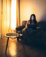 Young woman uses laptop watch course, checks email news online sitting on sofa. Beautiful girl working on a computer, watching a webinar at home in cozy blanket in winter by lamp photo