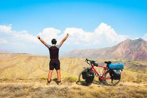 Close up back view inspirational excited joyful caucasian male cyclist on viewpoint by red touring bicycle in deserted mountains with hands up confident carefree lifestyle photo