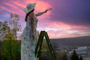 un mujer en un vestir soportes en un de madera escalera y pinturas el puesta de sol foto