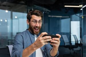 Mature businessman with beard is playing game on smartphone, man is having fun during break at workplace, businessman inside modern office. photo