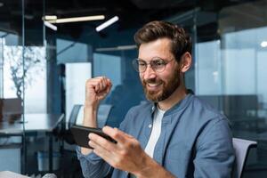 hombre dentro oficina a lugar de trabajo jugando juegos en teléfono inteligente, empresario contento con victorioso se regocija y sostiene mano arriba en triunfo gesto, jefe en camisa contento con logro y resultado. foto