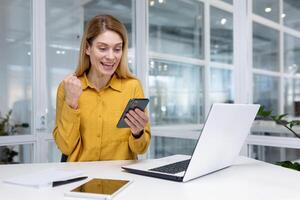 alegre exitoso negocio mujer celebrando victoria éxito, mujer jefe recibido en línea notificación en teléfono, financiero dentro oficina a lugar de trabajo, utilizando aplicación en teléfono inteligente foto