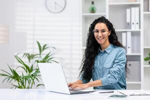 alegre Hispano mujer en un hogar oficina configuración, sonriente a el cámara mientras trabajando en su ordenador portátil con plantas y un reloj en el antecedentes. foto