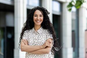 joven hermosa Hispano mujer con Rizado pelo sonriente y mirando a cámara, mujer de negocios con brazos cruzado fuera de oficina edificio al aire libre. foto