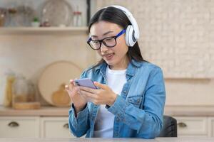 Young beautiful Asian woman listening to music on the phone. Sitting at home in the kitchen wearing white headphones, holding a smartphone, scrolling through the playlist, choosing a song, smiling photo