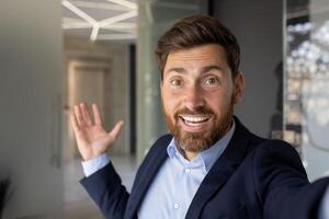 Close-up portrait of young smiling businessman standing in office indoors and talking on call, holding camera in hands. photo