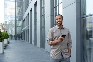 Hispano empresario caminando fuera de oficina edificio, hombre participación teléfono inteligente, sonriente y mirando a cámara foto