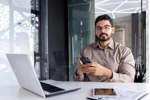 Portrait of successful Indian businessman, serious thinking man looking at camera at workplace inside office, boss holding phone, using laptop. photo