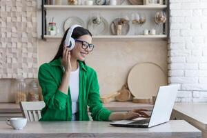 Young beautiful woman sitting in headphones at home at kitchen table and using laptop photo