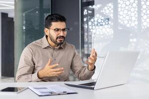 indio empresario mirando confuso mientras gesticulando durante un discusión a su moderno oficina. esta imagen capturas lugar de trabajo desafío y resolución de problemas escenarios. foto