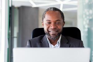 maduro adulto africano americano empresario en medio de oficina trabajando con computadora portátil, cerca arriba hombre sonriente mirando a computadora pantalla, satisfecho con logros jefe a lugar de trabajo cerca arriba. foto