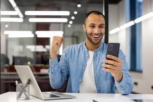 A cheerful young businessman in a light blue shirt is celebrating a victory, holding a smartphone at his modern office. The image evokes feelings of happiness, triumph, and professional success. photo