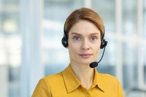 Web view camera, pov. Woman with a headset phone listens to the interlocutor, call, online meeting. The support service worker looks seriously and thoughtfully at camera, works inside office. photo