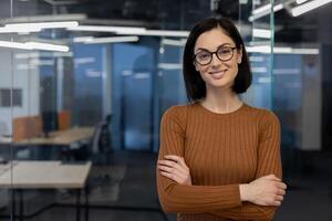 retrato de descuidado mujer en gafas y largo manga en pie con doblada brazos en antecedentes de vacío oficina. confidente morena mujer con corto Corte de pelo sensación satisfecho con construido carrera profesional. foto