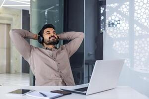 Businessman resting inside office with his hands behind his head with eyes closed dozing, man well done successfully completed a project with headphones listening to music and audio books podcasts. photo