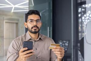 A focused man in glasses holds a credit card and smartphone in a modern indoor setting, possibly making an online payment. photo