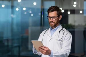 Mature and successful doctor in medical white coat and stethoscope working in boring clinic office, man in glasses smiling and using tablet computer, for online patient consultation. photo