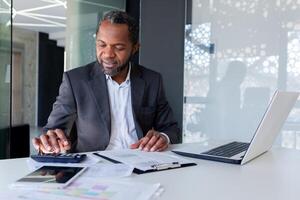 exitoso satisfecho financiero detrás papel trabajar, afro americano mayor experimentado relleno fuera documentos, utilizando calculadora para cálculo, empresario detrás papel trabajo dentro oficina sonriente. foto