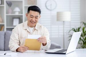 Happy and successful asian received mail notification envelope with good news, man sitting at table at home, working in home office in living room, joyfully celebrating winning news. photo