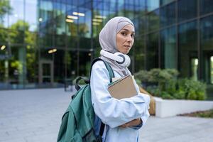 un joven musulmán mujer vistiendo un hiyab, auriculares, y que lleva un mochila y computadora portátil, soportes pensativamente fuera de un contemporáneo oficina edificio. foto