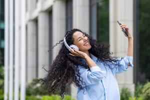 contento joven mujer utilizando teléfono mientras caminando en el ciudad de cerca, Hispano mujer en auriculares utilizando teléfono inteligente aplicación a escucha a música, alegremente canto y baile. foto