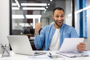 un indio hombre en un hogar oficina ajuste exuda felicidad mientras celebrando un negocio victoria, reflejando éxito y positividad foto