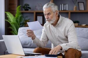 Focused senior man with gray beard sitting on a sofa, examining papers while working on a laptop in a modern home office setting. photo