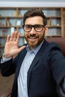 Mature businessman in glasses and suit smiling and waving at the camera, giving a friendly greeting from his cozy office. photo