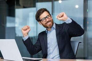 Mature businessman in office dancing sitting at desk, senior man working inside building celebrating successful achievement with laptop. photo