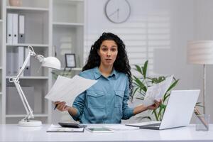 A Latina businesswoman looks confused while multitasking at her home office, surrounded by paperwork and a laptop. photo