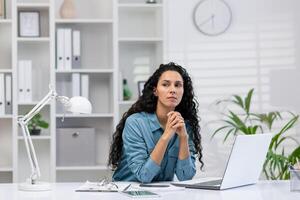 A serious hispanic woman works from her well-organized home office, demonstrating focus and professionalism in a peaceful environment. photo