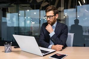 trabajo en el oficina de un financiero analista, banquero, empresario. un joven hombre se sienta a el mesa pensativamente a un ordenador portátil. foto