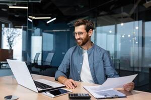 Mature businessman in shirt doing paperwork, man working with documents, contracts and bills sitting at table using laptop at work, financier accountant with beard and glasses. photo