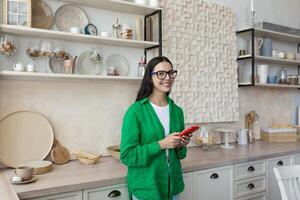 A young beautiful woman in glasses and a green shirt is standing in the kitchen at home,typing a message on the red phone,chatting with her friends,using a new application. He looks at the camera, smiles. photo