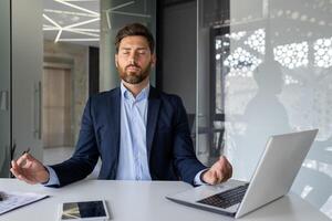 Mature successful senior experienced man meditating in lotus position inside office, businessman with closed eyes resting and visualizing future plans, boss in business suit at workplace. photo