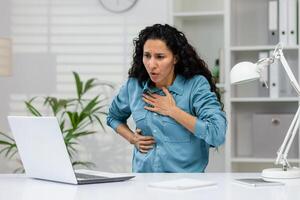 estresado mujer de negocios experimentando agudo cofre dolor, posiblemente un corazón ataque o ansiedad, mientras trabajando a su escritorio en un oficina ajuste. foto