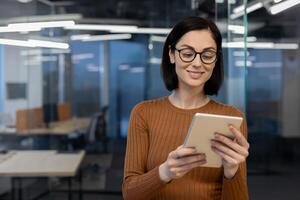 A content female professional in a brown sweater uses a tablet in a well-lit office space, embodying efficiency and comfort at work. photo