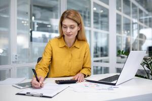 Serious thinking business woman behind paper work, woman boss concentrated writing documents working with papers graphs and charts inside office at workplace using calculator and laptop. photo