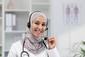 un alegre musulmán hembra médico en un hijab con un auriculares, Listo para un en línea consulta en un brillante médico oficina, mirando a el cámara. foto