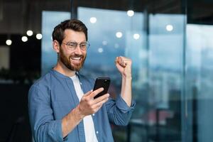 Businessman in shirt near window using phone , mature man with beard received notification online good news, worker celebrating victory rejoices, holding hand up gesture of triumph and achievement. photo
