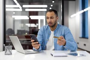 A man is sitting at a desk with a laptop and a cell phone. He is looking at his cell phone and he is in a serious or concerned mood photo