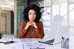 Serious diverse female boss strictly looking at camera while keeping chin on folded hands by table with clipboards. Millennial ceo putting computer aside and ready for discussing working moments. photo