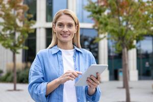 Smiling businesswoman in casual attire working on a tablet outside modern office buildings with natural foliage. photo