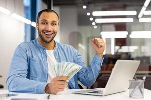 Joyful male entrepreneur with cash fist pumping in modern office, expressing success and excitement. photo