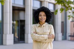 retrato de un joven hermosa africano americano mujer de negocios fuera de un oficina edificio, el mujer es sonriente y mirando a el cámara con su brazos cruzado. foto