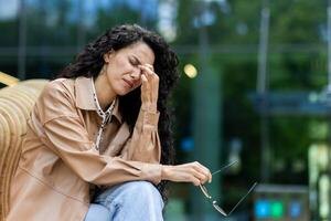 Sorrowful woman with luxurious black curly hair holding pair of glasses while touching face in nasal area. Despondent female sitting on outdoors bench and resting after hard work day on fresh air. photo