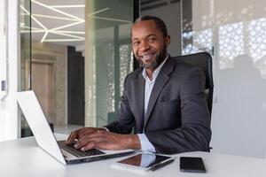 retrato de exitoso contento africano americano jefe, hombre sonriente y mirando a cámara, empresario en negocio traje sentado a escritorio con ordenador portátil dentro oficina. foto